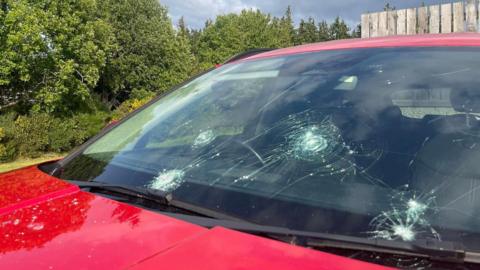The car's windscreen is smashed in several places. The car's bonnet is red.