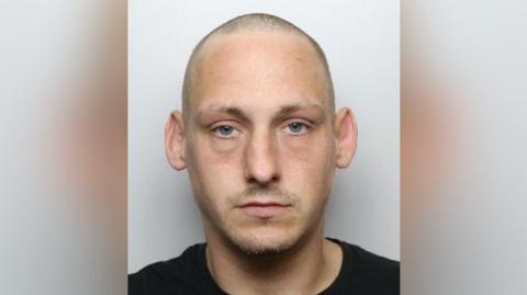 A mugshot of Corney, who has a shaven head and a little facial hair above his upper lip and along his chin. He is wearing a black top and looks straight at the camera