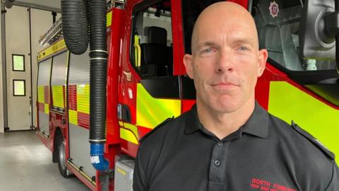 A man wearing a fire service polo shirt looks into the camera with a neutral expression on his face. He is standing in a large garage with a fire engine behind him.