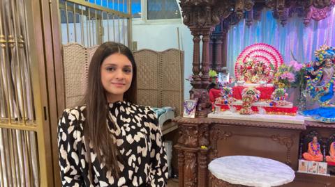 A woman with long brown hair wearing a black and white patterned top is next to a colourful shrine