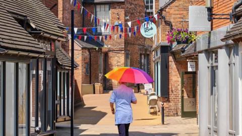 A woman walks up narrow road with shops on either side and holds a rainbow coloured umbrella over her head to shade the Sun