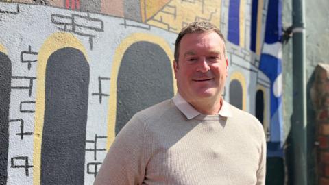 Head and shoulders image of Ryan Coogan standing in front of a colourful wall. He has short brown hair and is wearing a beige jumper with a light shirt underneath.