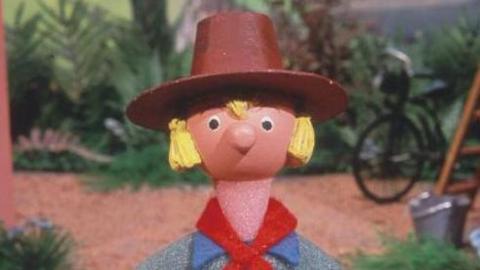 A small puppet with a pink face, long blonde hair, a brown rimmed hat, red neckerchief and blue shirt. The puppet is stood among plants and a brown background