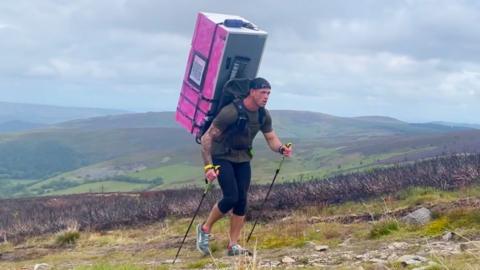 A man walks up a hill with walking poles, with a pink fridge attached to his back.