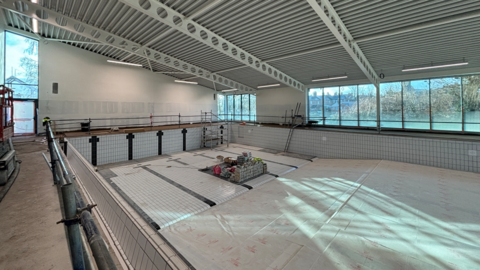 A partly-finished swimming pool with some of the tiles laid on the sides and floor. There are windows on the far side and a pile of building materials on the floor. 