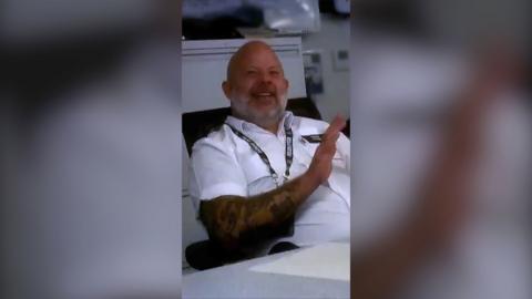 A police officer in white shirt, sitting back in a chair and smiling. He's bald with a beard.
