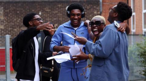 A group of four friends open their exam results and another classmate looks on in the background. They are smiling and and have their arms around one another. The student on the far left wears thick rimmed glasses and a black hoodie over a white t-shirt, and is facing the student to his right, who is wearing headphones and a blue tracksuit. Second from the right, another student with short blonde hair and sunglasses smiles towards the camera. Furthest on the right, a student in a blue shirt wearing glasses looks away from the camera towards the rest of the group.