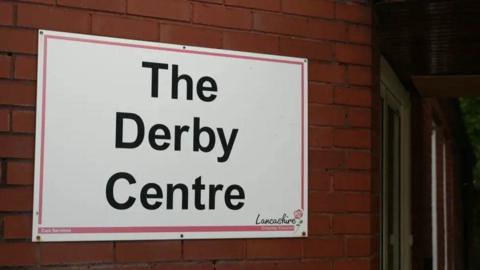 A white sign on a red brick wall, with the black writing saying, The Derby Day Centre, and Lancashire county council in the bottom right hand corner