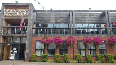 A public building with a wooden balcony above a brick ground floor, with pink hanging baskets above and bushes below the ground floor windows