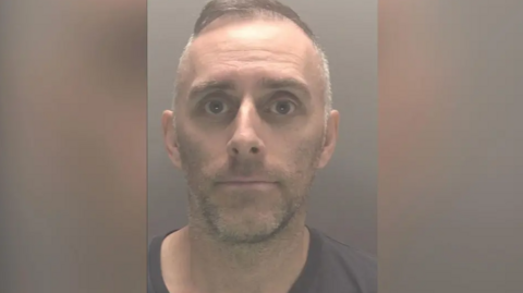 Police custody image of Scott Cameron. He has short grey hair and stubble wearing a brown t-shirt.