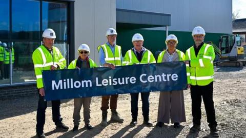 Six officials wearing white hard hats and hi-vis jackets hold a sign which reads, Millom Leisure Centre. They are standing in front of the facility, which is under construction.