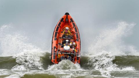 An RNLI lifeboat crashes through rough water. The orange RIB boat is riding the waves. It's bow is in the wave. Water is splashing into the area.