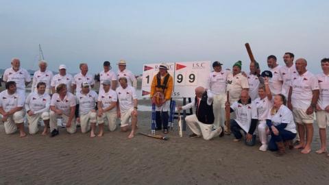 Two lines of players lined up on the sandbar mostly wearing cricket whites with a scoreboard and trophy in the centre 