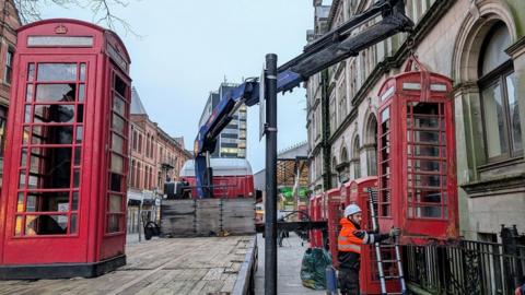Red telephone boxes are removed in Preston with a large blue crane by a workman in a high viz tangerine jacket and white hard hat. 