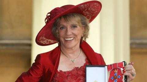 Dame Esther Rantzen holds a medal on the day of her investiture. She is wearing a red wide brimmed hat, a red dress, and is smiling. 