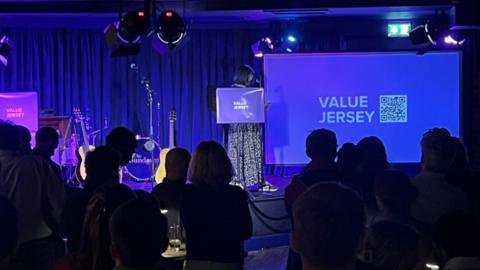 A woman is pictured speaking on a stage beside a screen which says 'Value Jersey' on it alongside a barcode. The woman has dark hair in a bob and she is wearing a long patterned dress. The backs of audience members heads can be seen in silhouette.