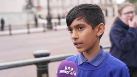 A boy wearing a blue jumper speaking into a Newsround microphone on the Mall