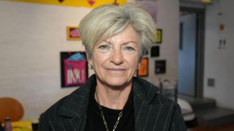 An older woman with short hair. She is wearing a black coat and top and has a gold chain necklace on.