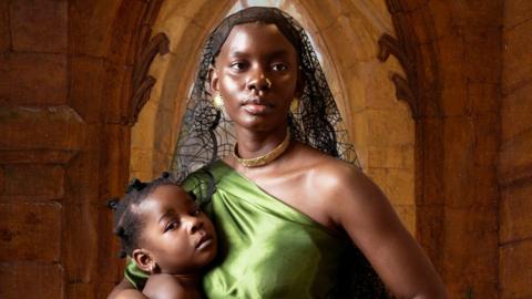 Dola Posh wrapped in a green cloth cradles her young daughter. A church arch way in the background frames her face.