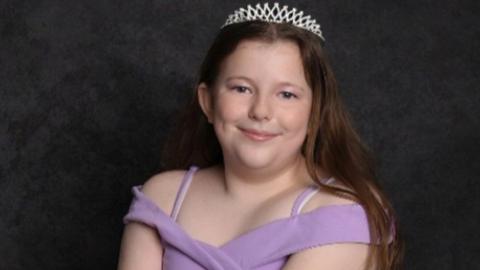 Leah Harrison, looking directly into the camera. She has long brown hair and pictured is wearing a silver tiara and purple dress.