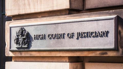 A metal sign on a sandstone block exterior wall that has a crest and the words: "High Court of Justiciary".