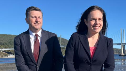 Douglas Alexander, wearing a navy suit and burgundy tie, sitting on a wall next to Kate Forbes who is wearing a dark blazer and pink top. Behind them is a bridge and water.
