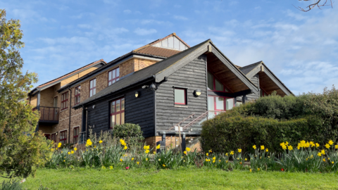 A landscape image of Farleigh Hospice's specialist inpatient unit in Chelmsford. It is a black wooden building in front of a brick building. It is surrounded by green bushes and grass and daffodils.