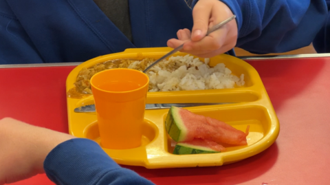 A yellow food tray with compartments for food is on a red table. There are two watermelon slices to the front right, a yellow up of water to the left, and in the main compartment at the back rice and chicken curry. There is a hand holding a fork scooping some of the carry, with a blue sweatshirt visible.