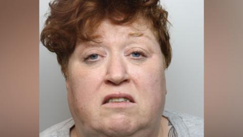 A police mugshot of a woman with curly auburn hair. She is wearing a grey top. She is positioned against a light-coloured backdrop.
