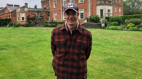 Harry Grafton, wearing a cap, and a red and black checked jacket, looking at the camera and smiling, standing on a grass lawn, with a large red brick building behind him, that looks like a stately home. There are steps to the building behind him and ornate garden features. 