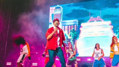 A picture of the actor and rapper Will Smith on the Open Air Theatre stage wearing a red shirt and blue jeans and surrounded by dancers.
