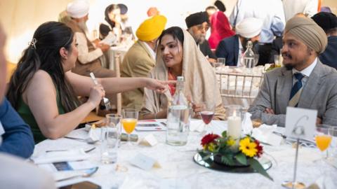 People sit around a large table with a white tablecloth and centre setting of candle and red and yellow flowers in a marquee, with drinks in front of them. A woman with long black hair is talking to a woman in a head covering. A man in a suit and turban looks on. Others can be seen at tables behind, some wearing turbans and head coverings.