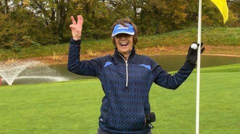 Lyn Parry holds up three fingers and a flag to celebrate her hole-in-one treble success
