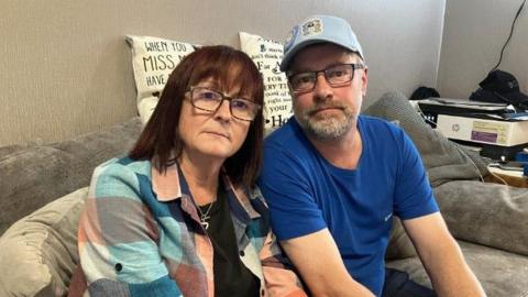 A man and a woman sat on a grey sofa, looking at the camera with neutral faces. Propped on top of the sofa are two memorial cushions for their son.