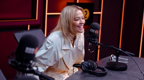 Artist Rita Ora smiling and laughing in a Radio 2 recording studio. She is wearing a white coat and next to a microphone and headphones