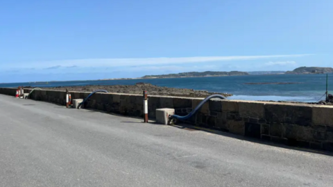 Black Rock - A site at the North of the Bridge with blue sea, looking out to Herm. In the fooreground are pipes going into the sea.