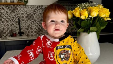 Baby Beatrice sits atop a kitchen counter wearing a half red and half yellow baby gro. Each side has the respective badges on. She has short red hair and is smiling slightly at the camera, with her hands outstretched. There is a vase of yellow flowers behind her.