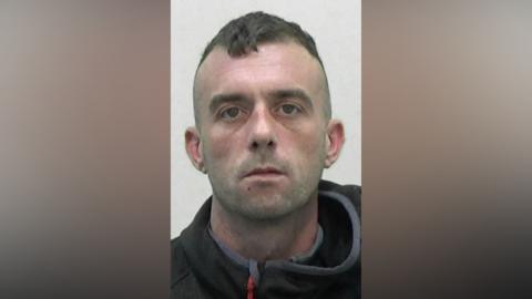 Mugshot of Toward. He has faint stubble and short dark hair on top in a drooped quiff with the sides shaved. He has a vacant look in his eyes.