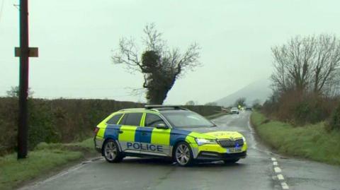 A police car parked across a rural road at a T junction. There are bushes and trees on either side of the road and the sky above is grey. 