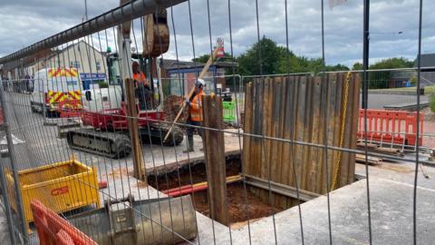 Roadworks site with fenced-off excavation, workers in high-visibility clothing using an excavator beside a dug trench, with barriers, traffic management cones, and a service vehicle parked nearby under a cloudy sky.