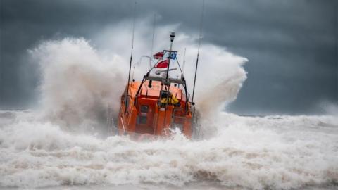 An orange RNLI lifeboat seen from the back launches head-on into heavy seas, causing a huge bow wave to spray up at either side of the vessel. White foaming waves crash in the foreground and a dark, stormy sky forms the background.