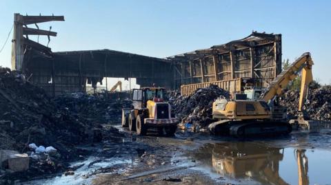 The aftermath of the fire shows a burnt out warehouse and materials