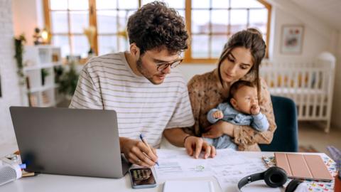 Stock photo shows a man and woman at home looking at bills and a laptop with a young baby