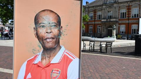 A large photo of a black man wearing a red and white Arsenal football shirt, who is part of the exhibition, with his face scratched and etched out. 