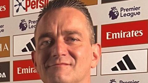 A man, with brown hair, smiling for camera. He is in front of a board full of adverts.