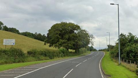 A71 east of Newmilns. The road curves round a corner in the distance and has the word "SLOW" on the road. Trees and bushes line the road and a field is seen on one side. 