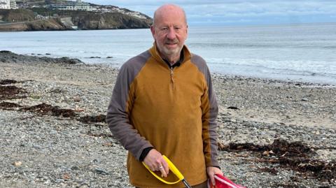 Bill Dale, a man with a grey beard, wears a brown and grey fleece and smiles. He is on the beach and you can see the headland in the background.