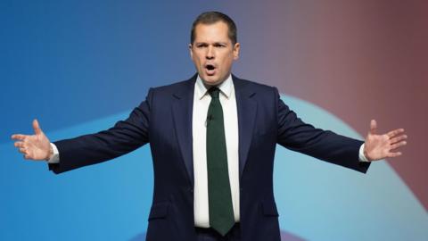 Robert Jenrick wearing a blue suit, delivering a speech on stage at the Conservative Party conference in Birmingham