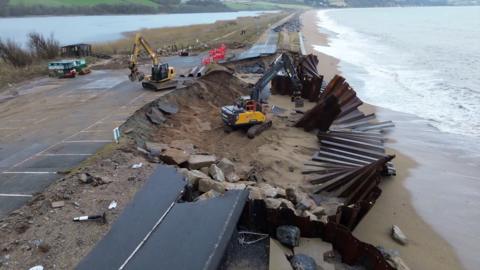 An aerial view of a scenic coastal road in Torcross, Devon that runs out along a shingle bar.  There is a large section that has been washed away and you can see where the road has collapsed. The road is closed as engineering work to shore up the road continues