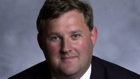 The image shows Oliver Colvile dressed formally in a dark suit, white shirt, and patterned tie, standing against a plain, light-colored background.